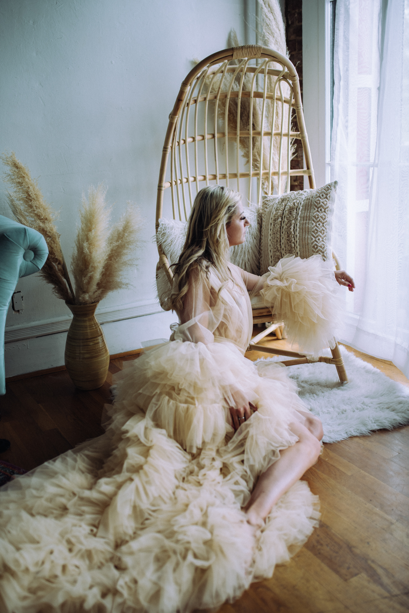 woman in beautiful tulle dress sits in a chair and stares out the window during her photo session