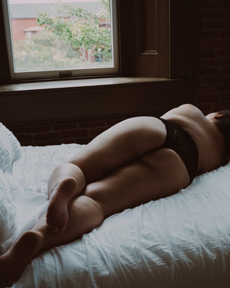 woman laying on a bed staring out the window in black underwear during an intimate portrait photography session