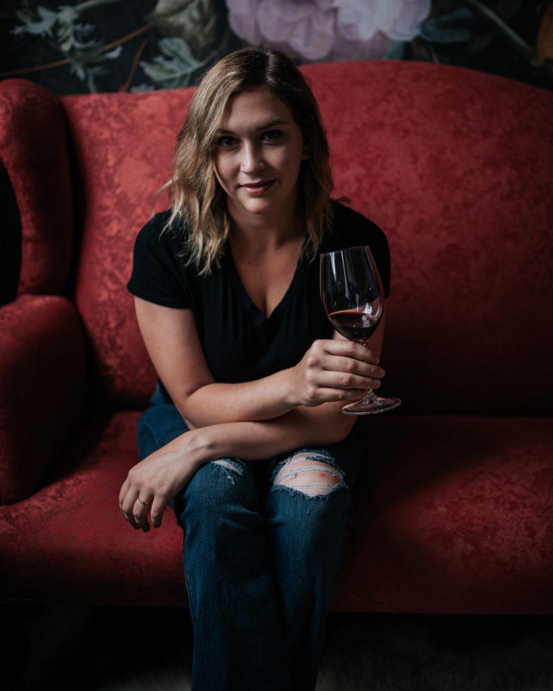 woman sits on a red couch with a glass of wine posing during her luxe headshot session