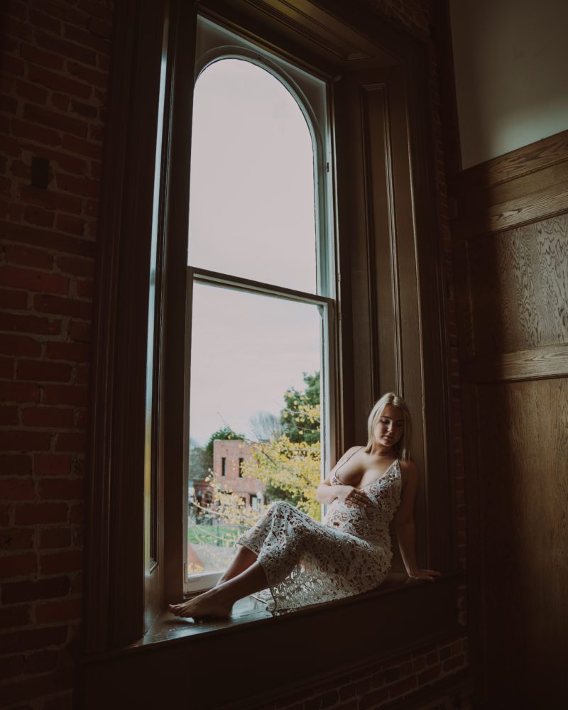 woman sits next to window during her maternity boudoir session posing for a photo in a white dress