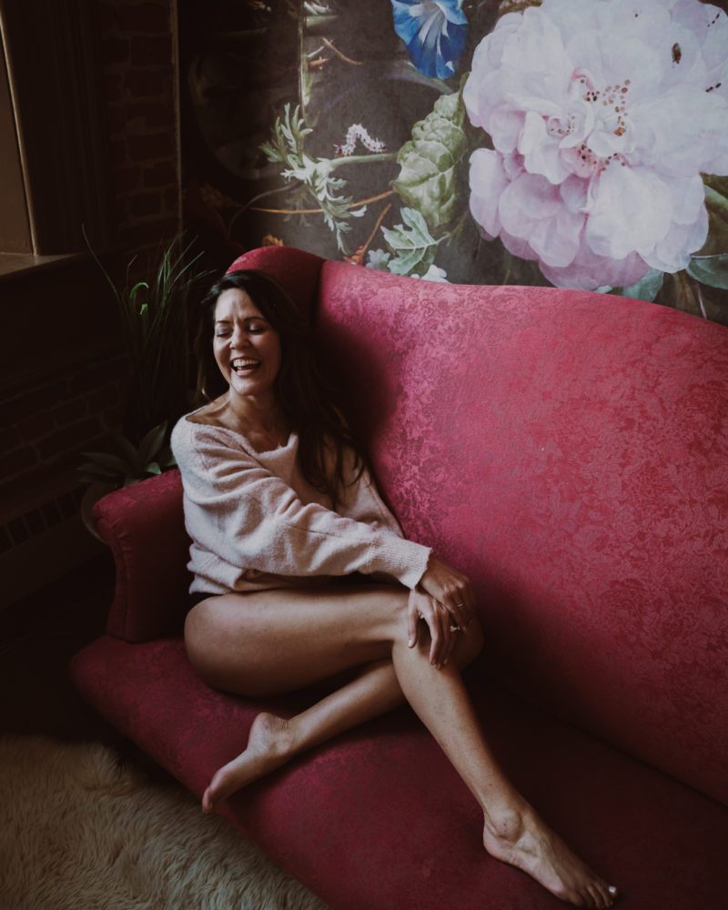 woman laughing while sitting on a red couch during her empowering intimate portrait experience with Kimberly Ann Boudoir in Humboldt County